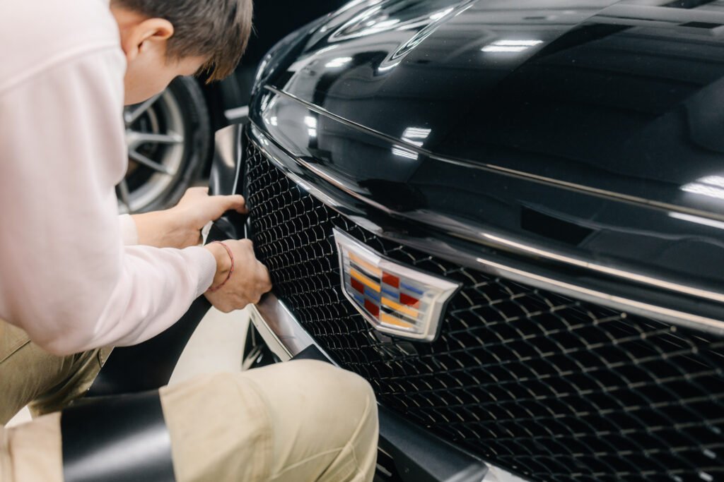 Technician carefully installing or adjusting the front grille on a black Cadillac inside a detailing studio.
