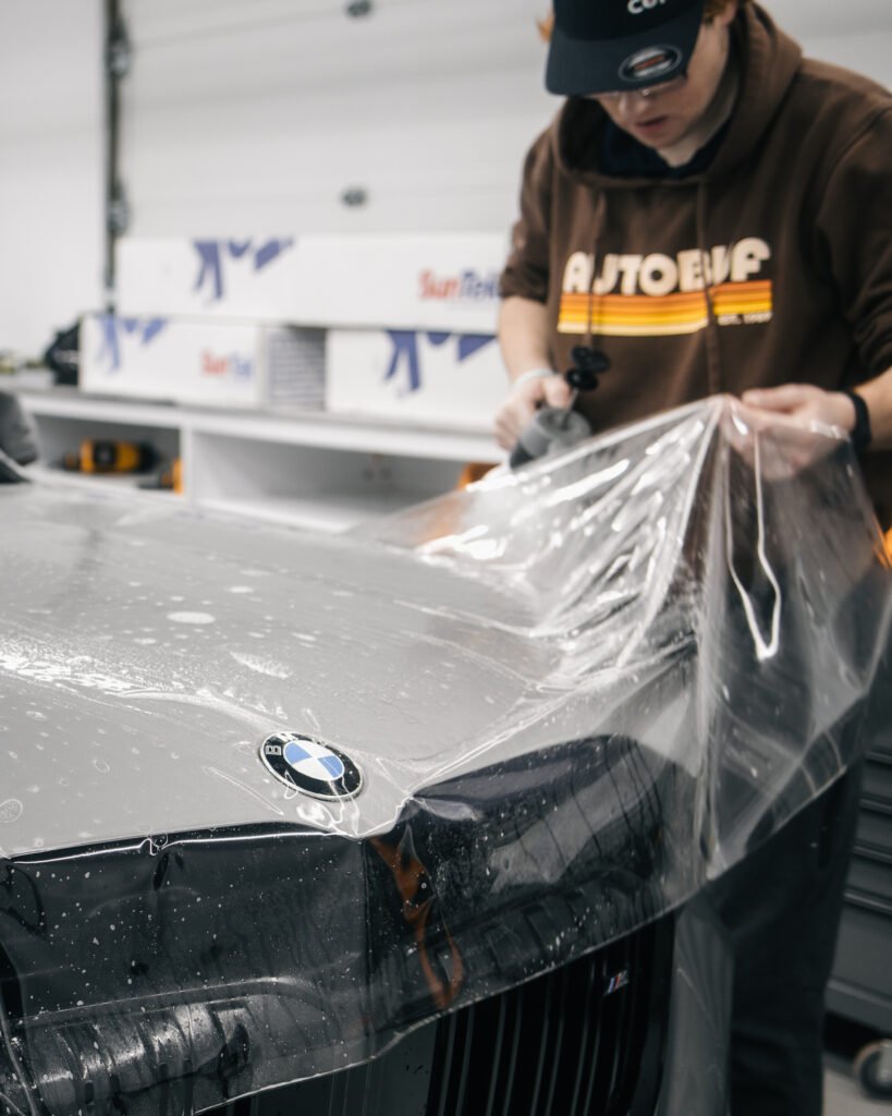 Automotive technician installing paint protection film on the front of a BMW inside a detailing studio.