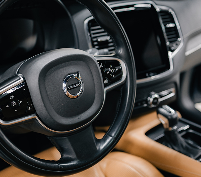 Close-up of a Volvo steering wheel and dashboard interior, featuring the Volvo logo, leather trim, and center infotainment controls.