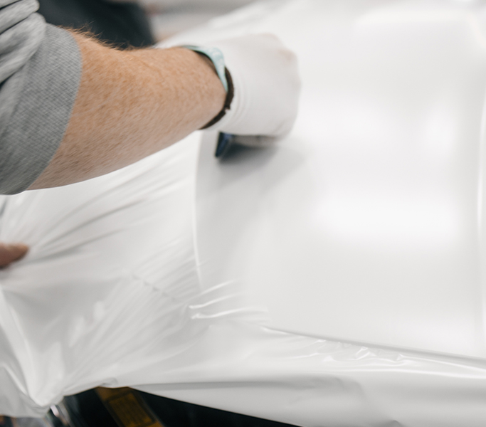 Technician applying paint protection film to a white vehicle panel, smoothing the clear film by hand for a precise fit.